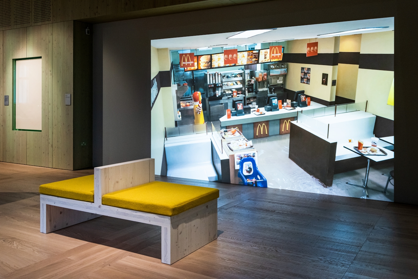 Photograph of a gallery installation showing a bench with yellow cushions facing a large video projection. The projection shows the inside of a McDonalds restaurant looking down from a high viewpoint in which the restaurant floor is beginning to flood with water.