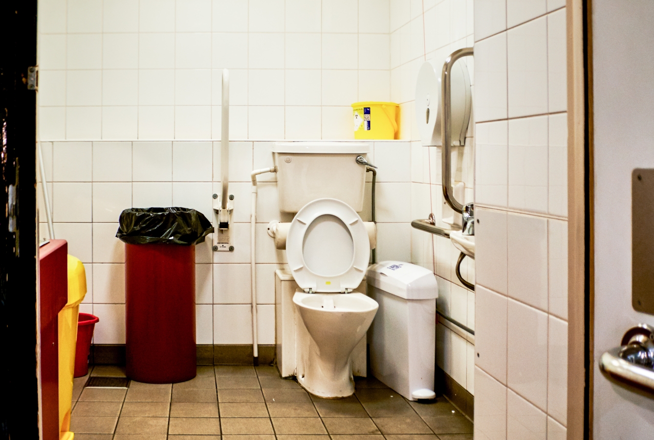Photograph though the doorway of an accessible toilet showing part of the door and doorframe, along with the contents of the room, toilet, basin and sanitary bins. The room looks run down and dirty.