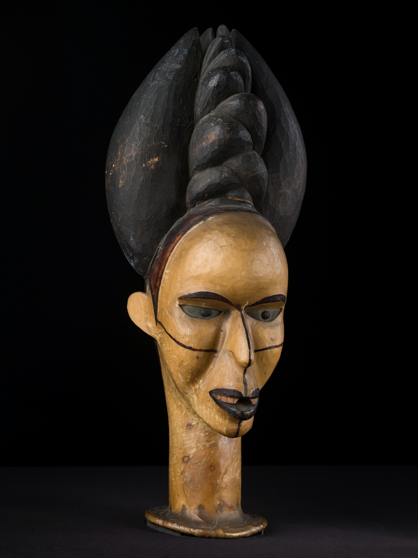 Photograph of the carved wooden head of a woman against a black background. On the woman's head is an elaborate headdress. Across her face thin black lines are drawn from the top of her nose to her chin and from ear to ear across her cheeks.
