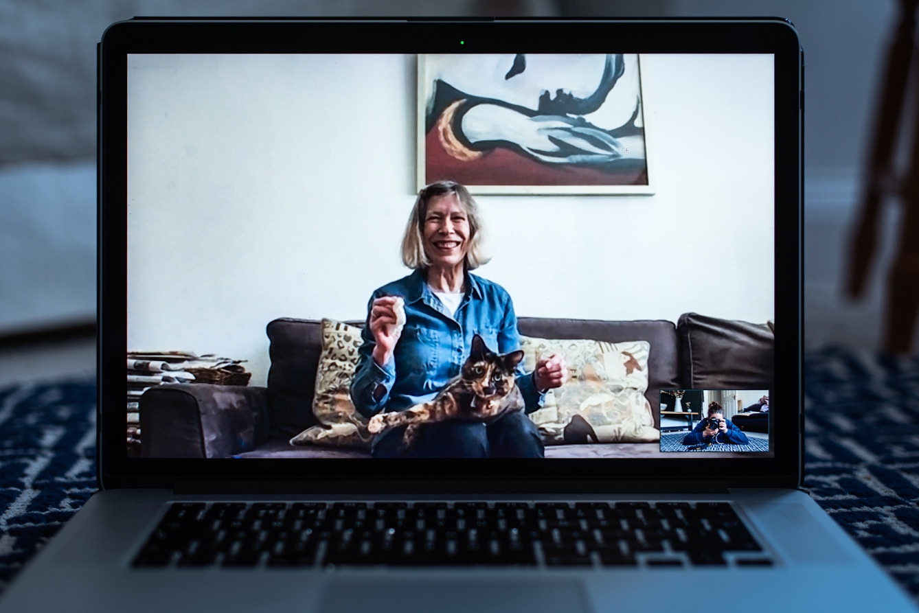 Photograph of an open laptop within a domestic scene. Most of the image is taken up with the screen, with part of the keyboard and trackpad visible. On the screen is a video call showing a woman sitting on a sofa with a cat on her lap. In the bottom right corner of the screen the photographer can be seen in a small floating window, camera to her eye, in the process of taking the picture.