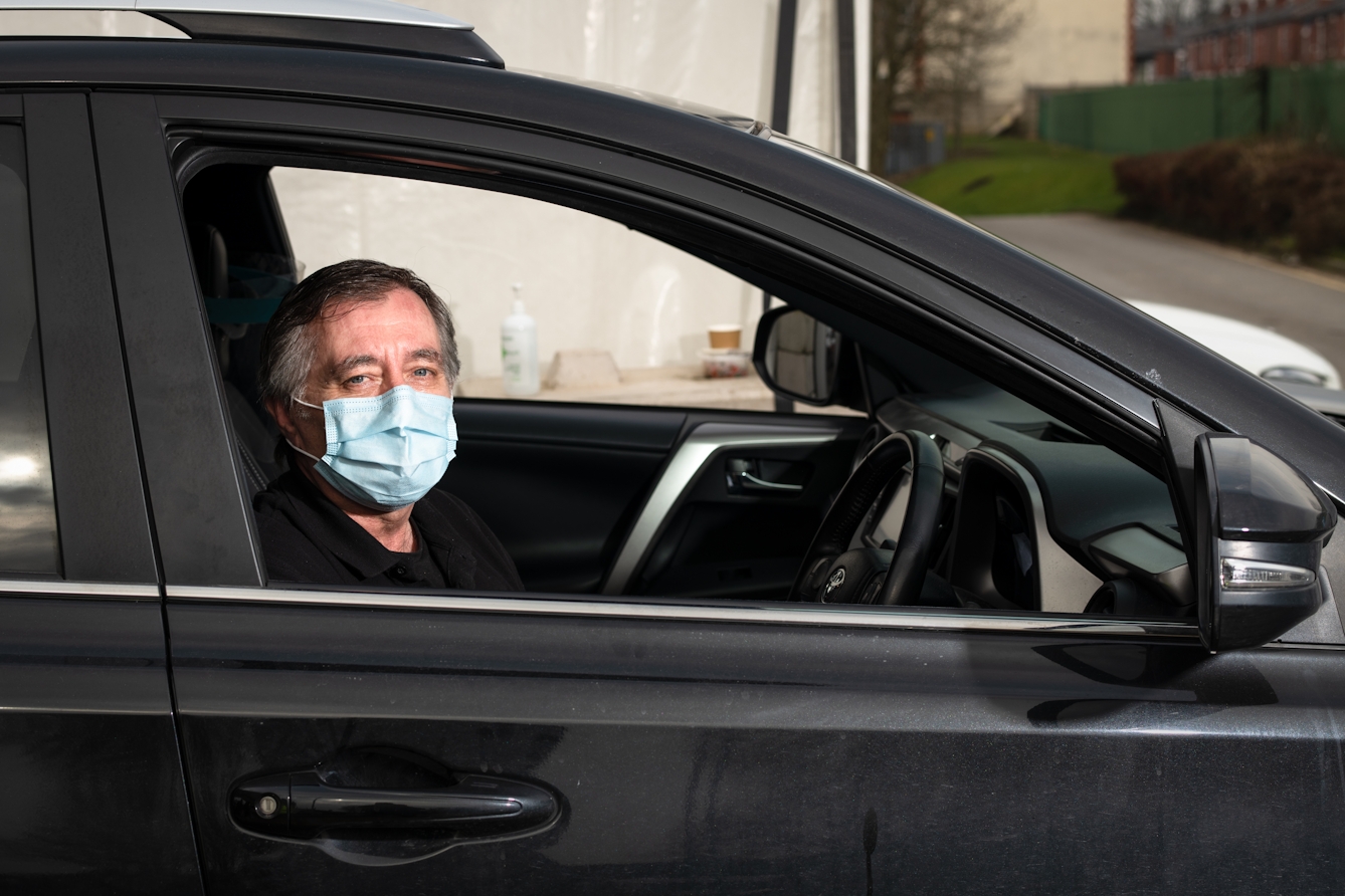 Photographic flash lit portrait through the open side window of a stationary car. The car is black and the front of the car is facing to the right. Sat in the driver's seat is a man, looking to the camera. He is wearing a blue medical face covering. In the background is the out of focus side of a white marquee and distant buildings and lawn.