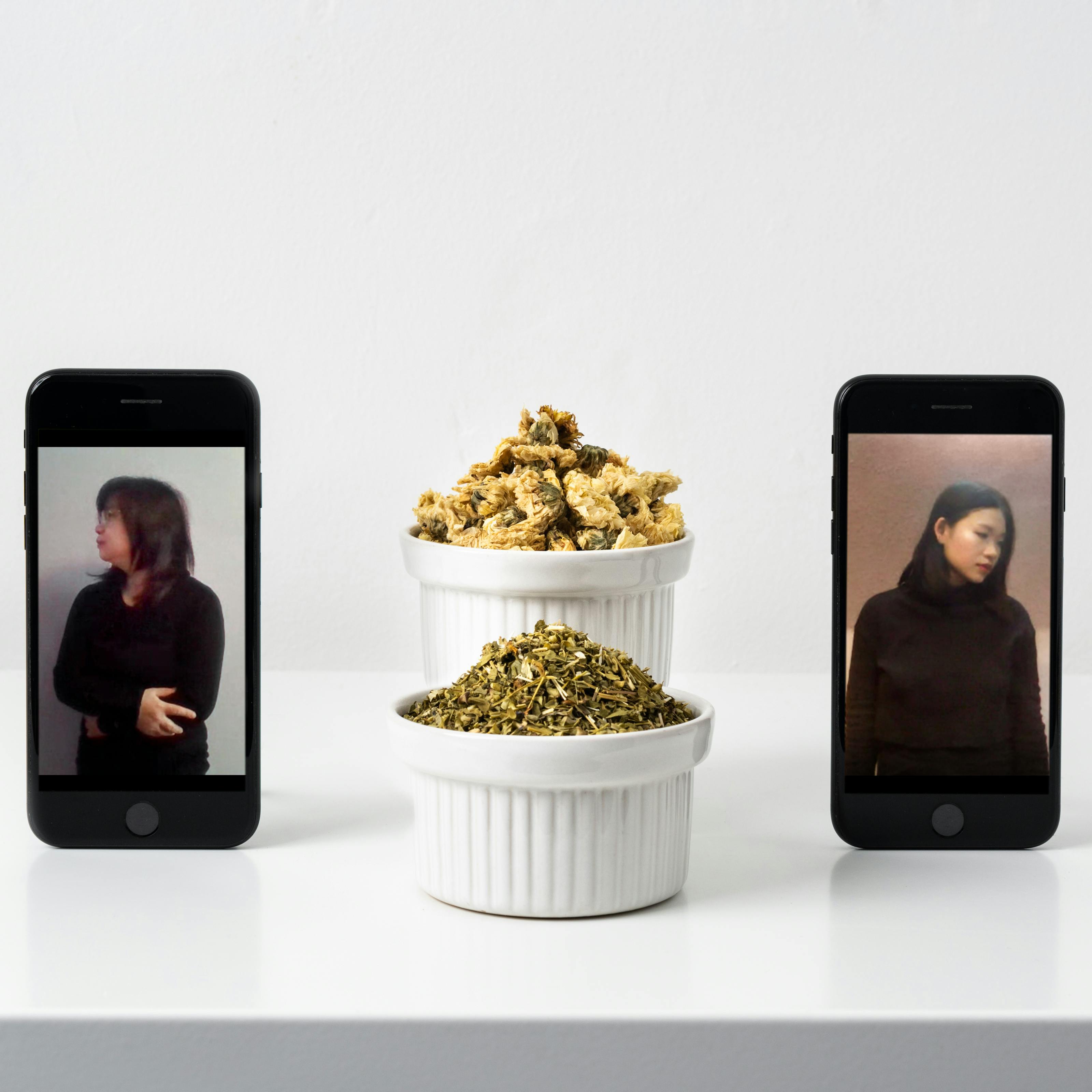 Photograph of a white, slightly glossy self against a white wall. On the shelf standing upright are two smartphones. On the screen on the left hand phone is a portrait of an older woman dressed in black from the waist up looking off to the left. On the screen on the right hand phone is a portrait of a younger woman dressed in black from the waist up looking off to the right. Between the two phones are two white ramekins one behind the other, containing dried Chrysanthemums in one and dried Angelia in the other.