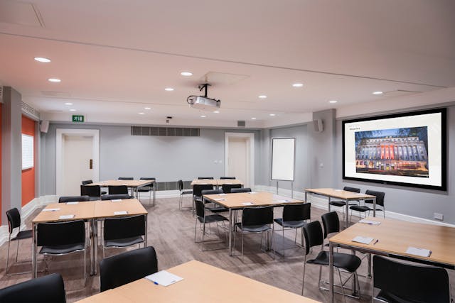 Photograph of the Burroughs room at the Wellcome Collection. 

Photograph shows a cabaret set-up, with desks placed in small groups, and a presenting screen and whiteboard at the front of the room.
