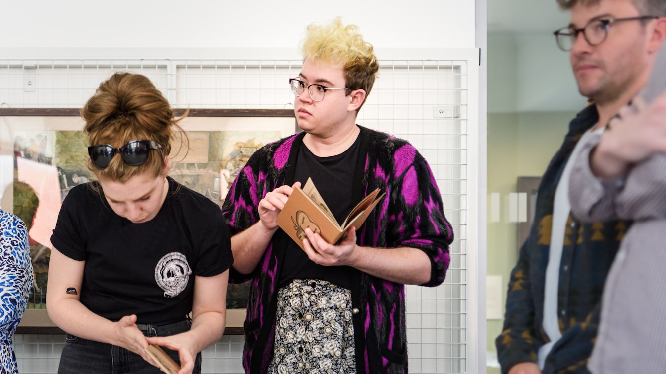 Photograph of a group of individuals standing in a library room looking at collection materials. In the centre of the image an individual listens to a speaker (off camera) whilst carefully holding a small book in their hands. To the left an individual bends forward looking at a smaller book held in their hands. To the right more individuals are looking at and listening to the speaker, In the background library racking is bolted to the wall and a framed artwork hangs from it.