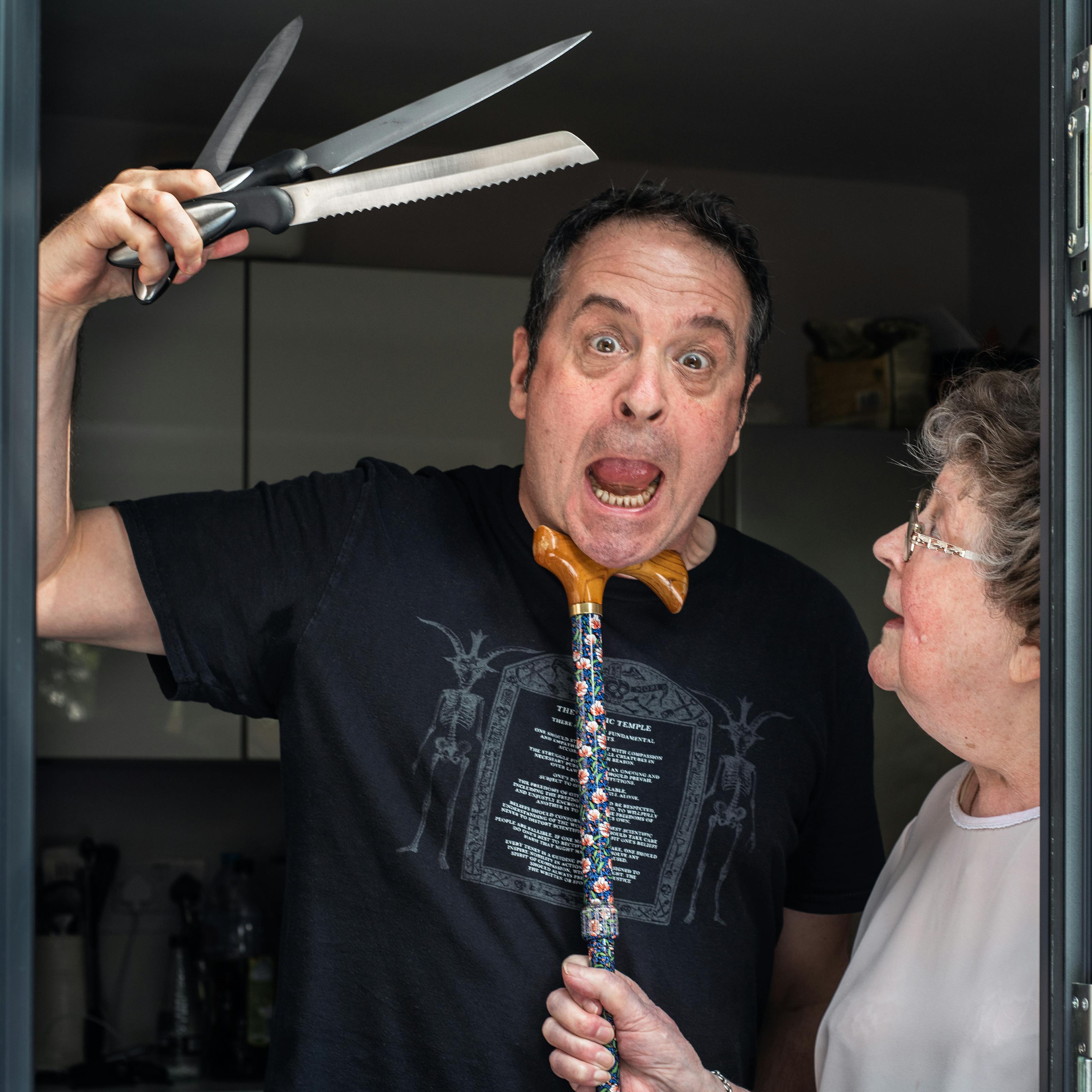 Photograph of a man standing in a glass patio doorway with his mother. The man is looking straight to camera with his mouth open, eyes wide and slightly mad look on his face. In his right hand held above his head are 3 kitchen knives held in a fan shape. His mother is standing to his left in profile, looking up at his face. In her right hand she is holding a walking stick, the handle of which she has tucked under his chin in a disciplinary fashion.