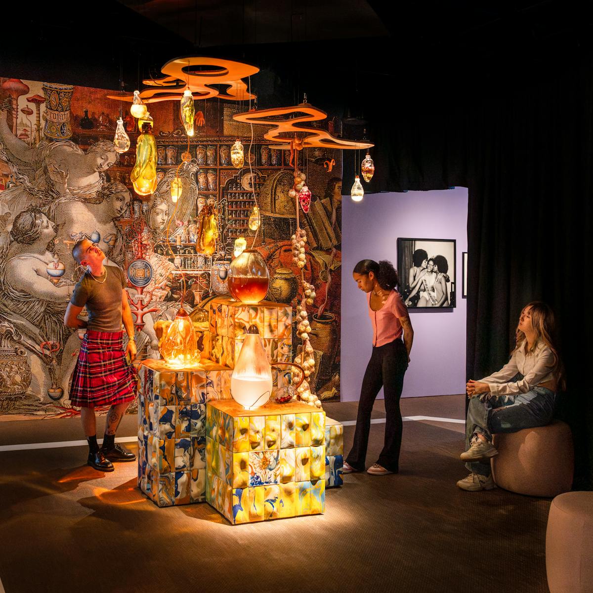 Photograph of 3 gallery visitors looking at an installation. At the centre is multi-levelled podium covered in ornate coloured tiles. Resting on the defaces and suspended above are organically shaped glass vessels containing coloured liquid and natural substances. The wall behind in covered in renaissance images. The whole scene is dark but warmly lit.
