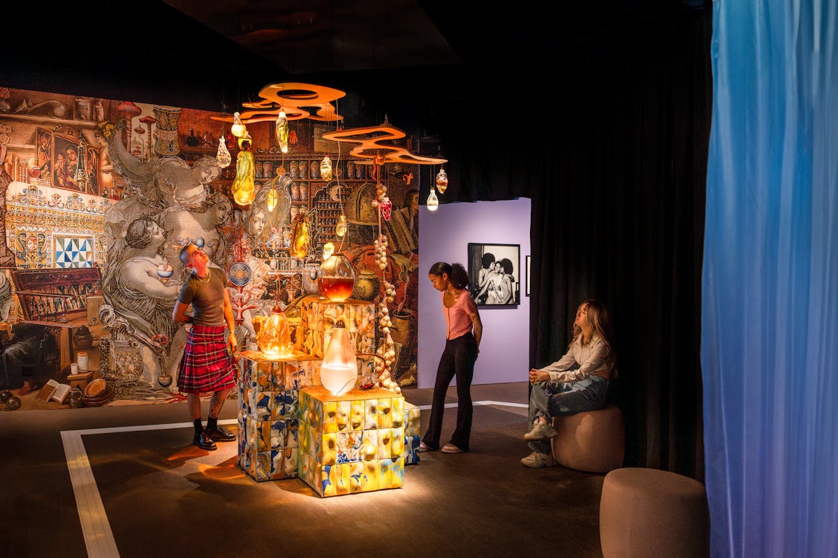 Photograph of 3 gallery visitors looking at an installation. At the centre is multi-levelled podium covered in ornate coloured tiles. Resting on the defaces and suspended above are organically shaped glass vessels containing coloured liquid and natural substances. The wall behind in covered in renaissance images. The whole scene is dark but warmly lit.