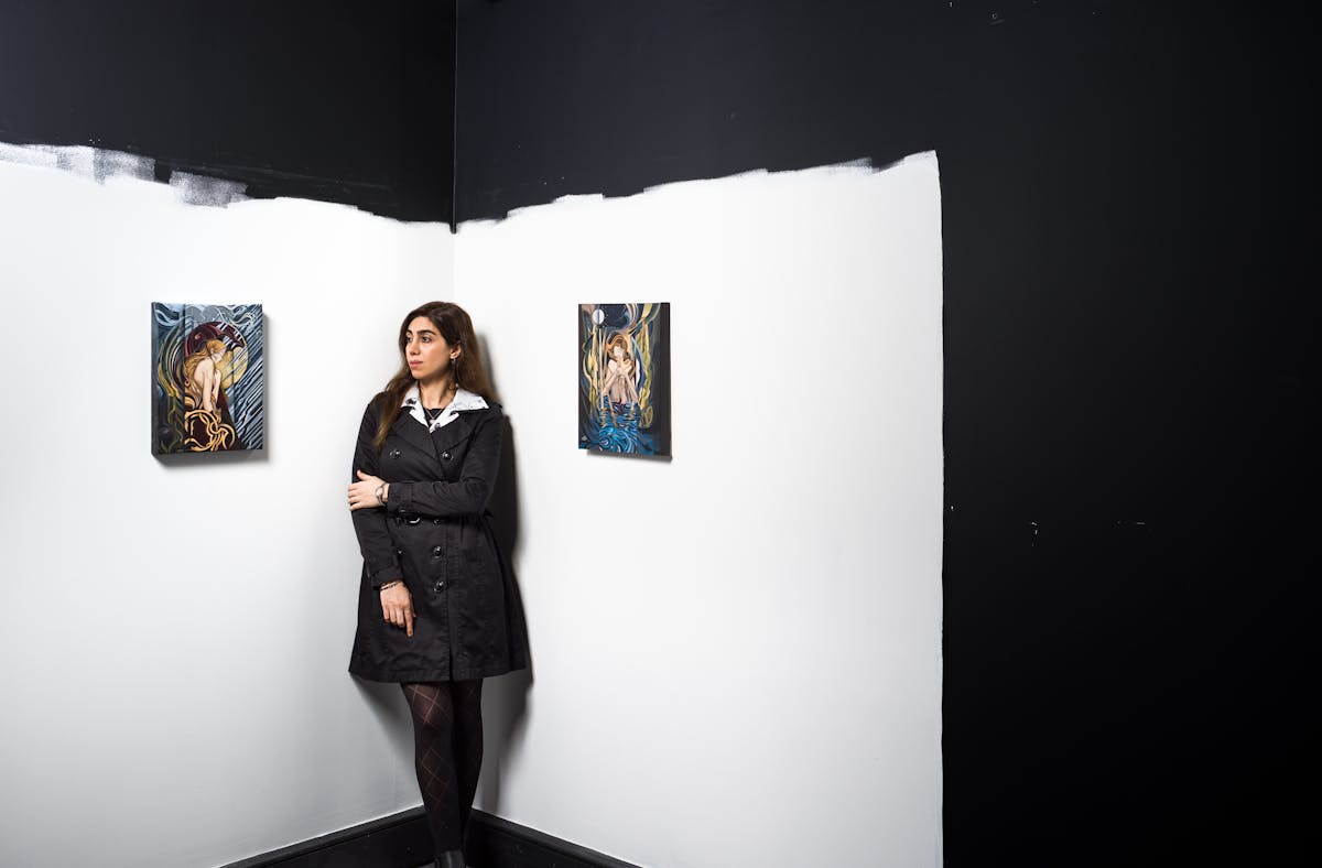Photograph of a young woman in a black dress with white collar, leaning into the corner of a white walled room. She is gazing off to the left. Hung on the walls to her left and right are two colourful paintings. Above her the white of the wall stops in a horizontal line of paint roller marks, giving way to a jet black wall. To the right of the image the white of the wall again stops abruptly, giving way to more black wall.