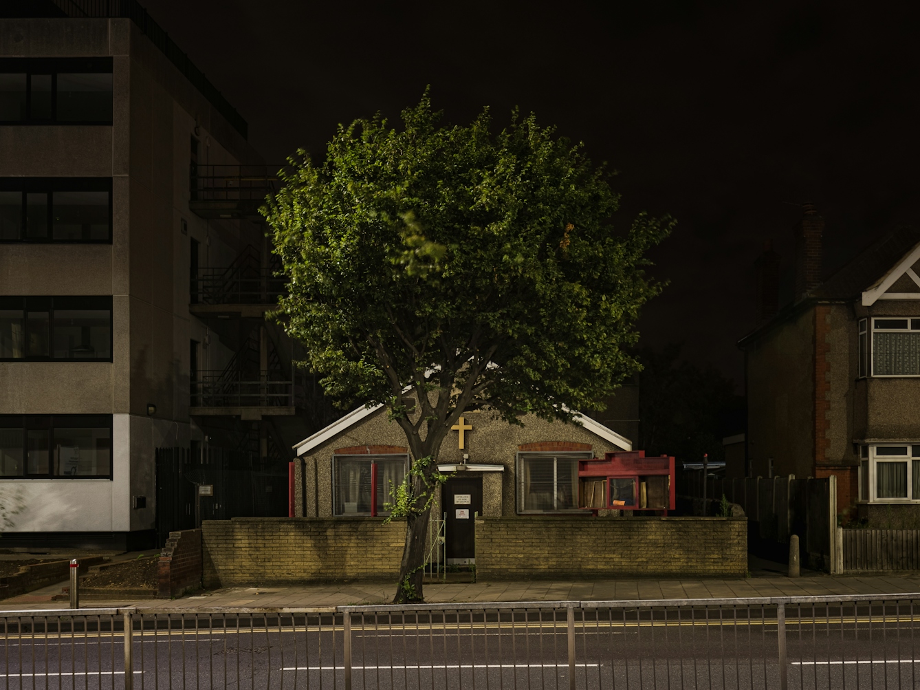 Photograph of Romford Christian Spiritualist Church at night. The single storey church has red and white features with a pitched roof.  The centre of the facade shows a gilded cross, with the facade itself being pebbledash and is partly hidden by a tree.  To the left of the church is a multi-storey commercial office, and to the right is a double storey post-war residential building. 