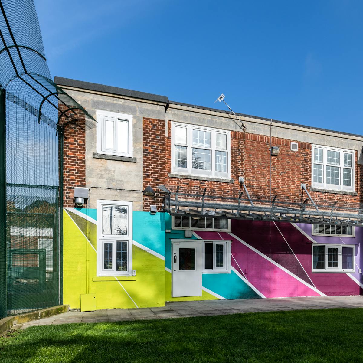 The photograph shows a flat roofed two-storey brick building with white PVC windows surrounded by a tall perimeter fence with a grassy area in the foreground. The lower storey of the building is painted with wide brightly coloured diagonal stripes in lime green, sky blue and various tones of purple separated with narrow bands of white. Under the windows on the upper storey there appears to be a safety fence.