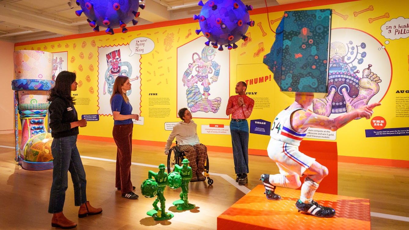 Photograph of a colourful exhibition space with four individuals facing each other speaking, next to a sculpture of a man in sportswear with a television instead of a head. On the wall in the background is a bright yellow mural containing drawings and text. 
