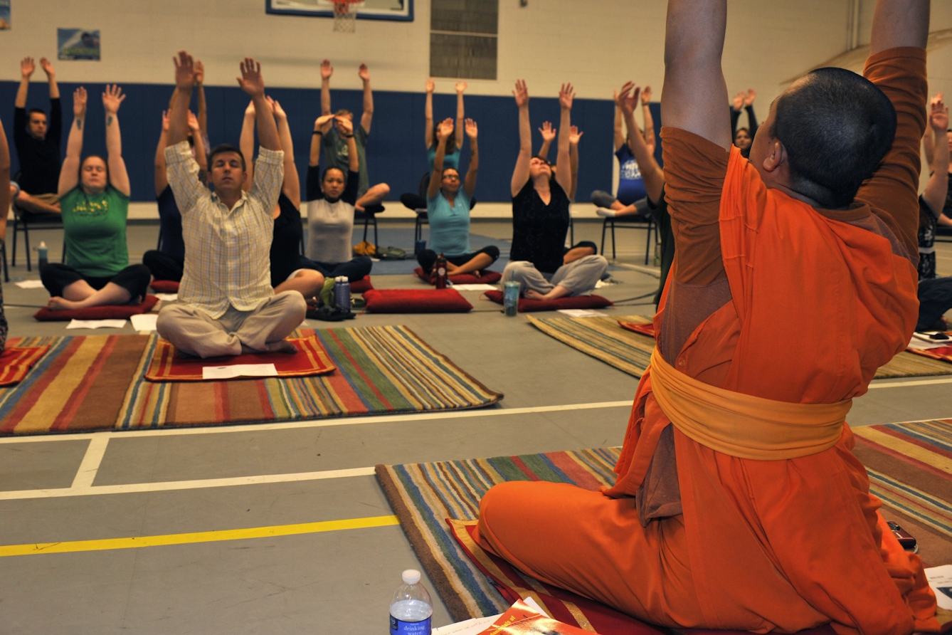 A person in orange robes sitting on a mat with arms raised in front of a group of people in casual clothing copying his actions.