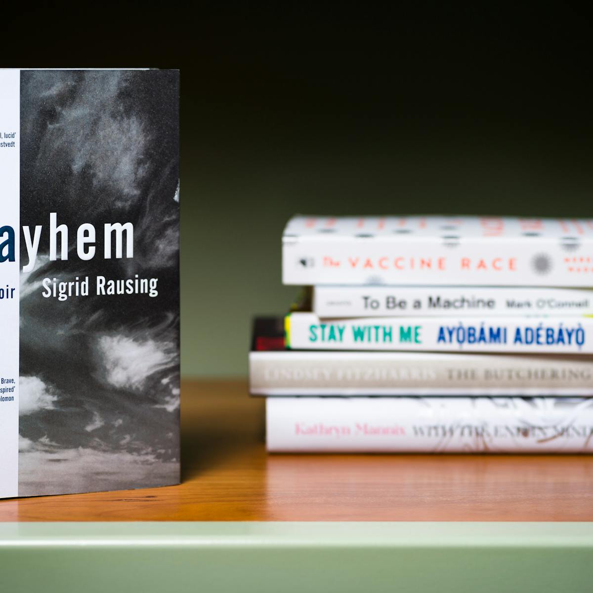 Photograph of a a stack of 5 books lying horizontally on a shelf with another book standing vertically in the foreground.