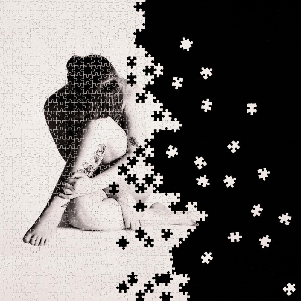 Photograph of a part made jigsaw puzzle containing hundreds of pieces. The jigsaw has been photographed against a black background, with many of the unused pieces scattered around in the black space. The image on the jigsaw is a warmed toned, monotone photograph of a nude woman against a plain light background. The woman is seated on the floor with her legs and arm crossed across her body, her head resting on her left knee such that her face cannot be seen. On her arm and leg are tattoos. The missing jigsaw pieces create a black void which encroaches from the right side of the image, fragmenting the image of the woman.