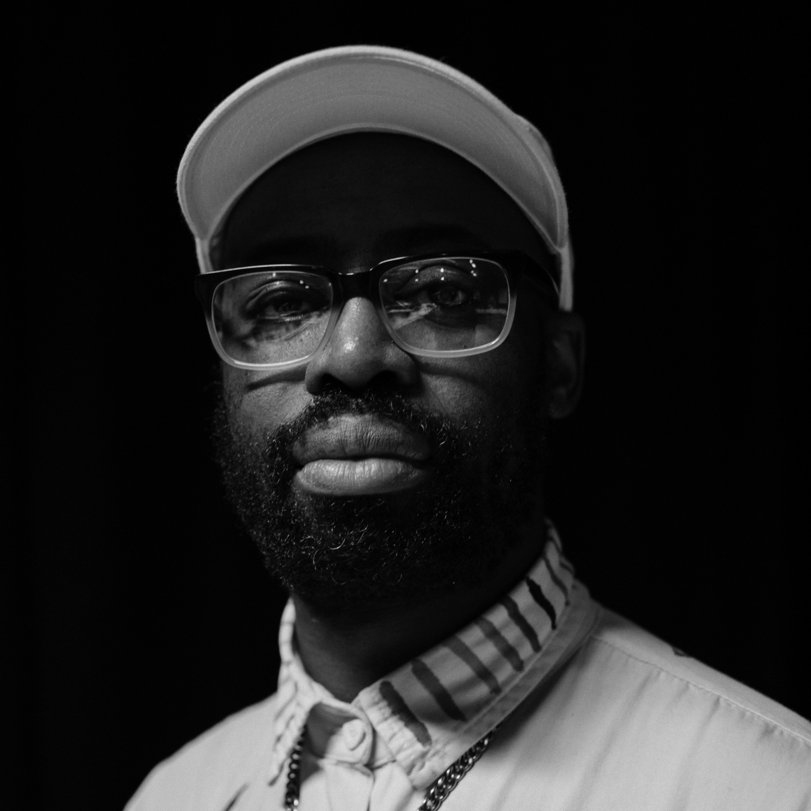 Black and white photograph of a young man with 
a short beard wearing glasses and a white baseball hat. The background is black.