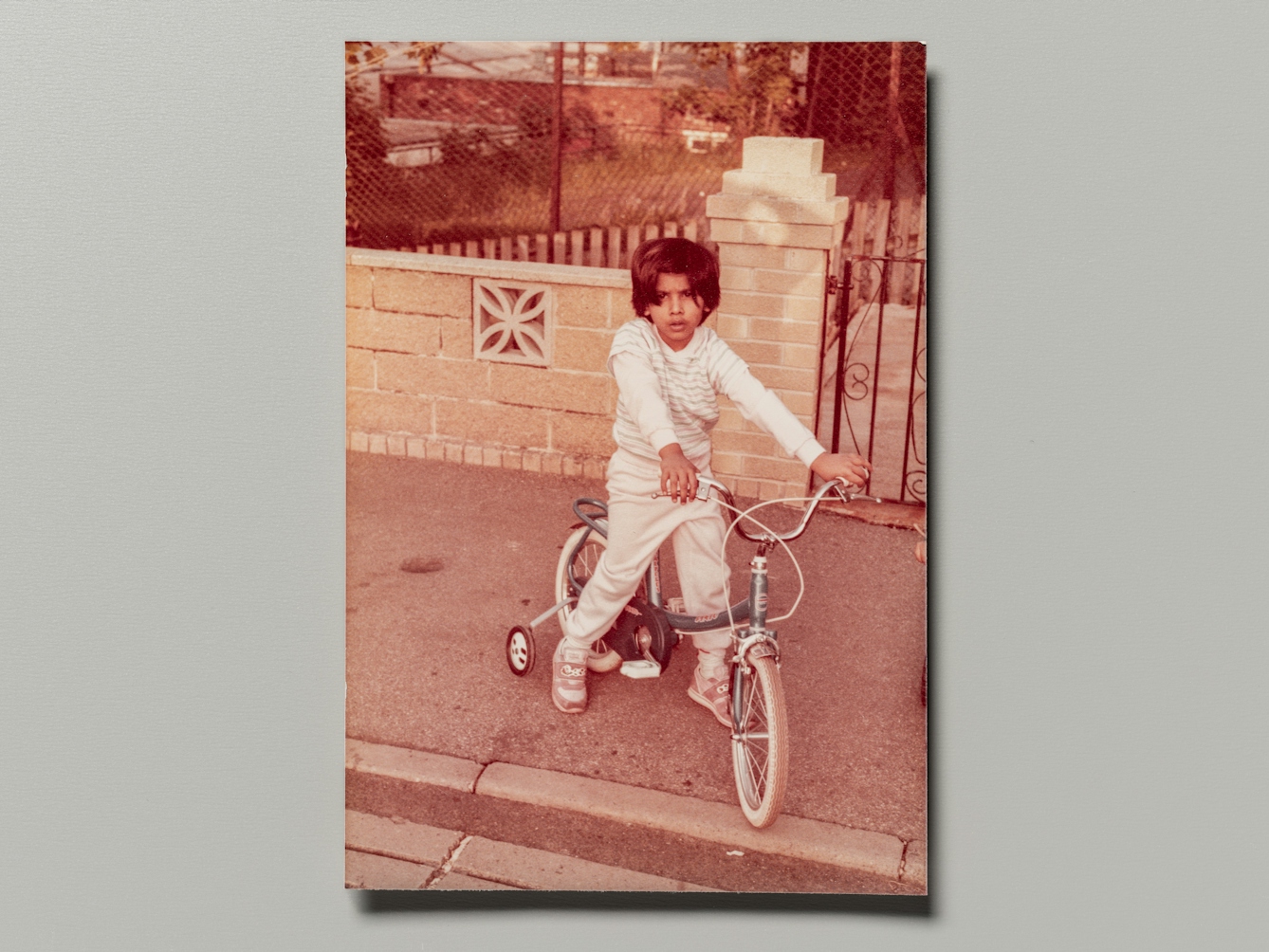 Photograph of a family photographic print resting on a grey background. The photo shows a young girl sat on a bicycle equipped with stabilisers, looking to camera. She is on the pavement of a residential street.