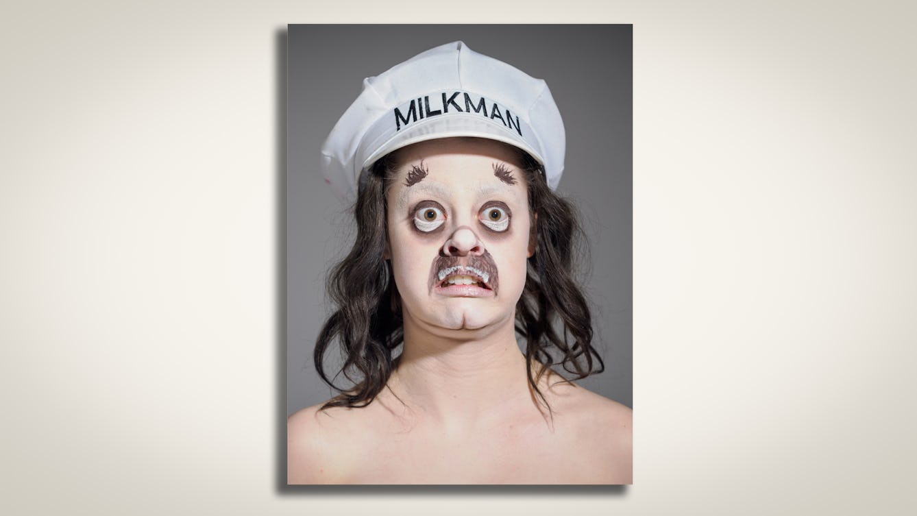 Head and shoulders portrait of drag artist Dairy King who has a shocked expression, a drawn on brown moustache and a white milk moustache who is also wearing a large white hat with the words "MILKMAN" printed on it. 