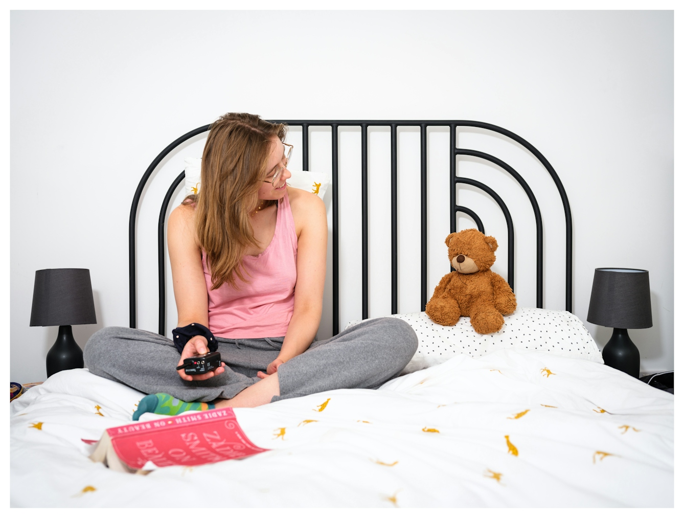 Assisted Self-Portrait of a woman sat cross-legged on her bed smiling across at a teddy bear sat the pillow next to her. In front of her on the bed is an open book, pages down. In her right hand is the remote trigger she's used to capture the image.