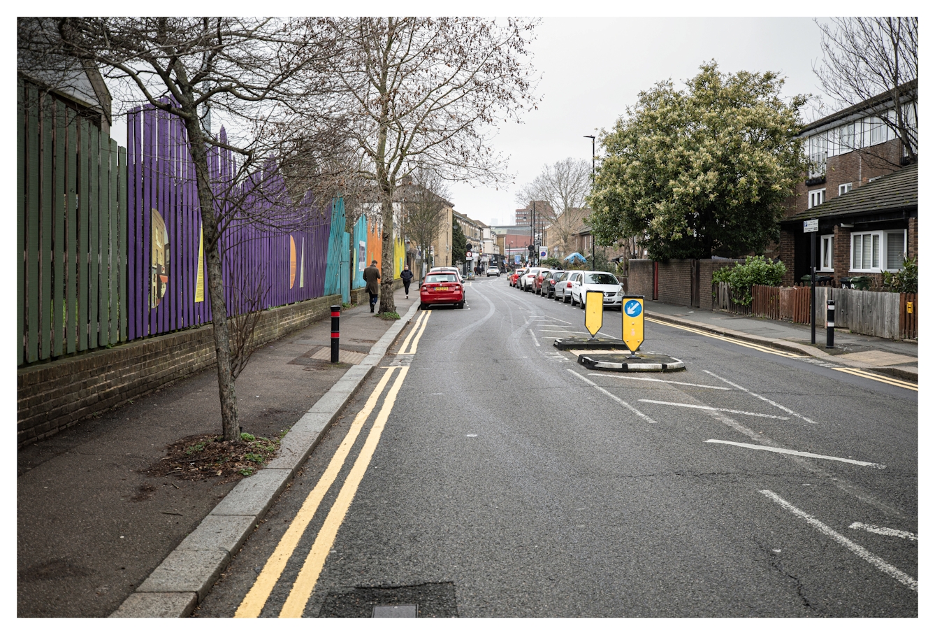 Photograph of a South London street scene in Brixton, showing a straight road receding into the distance. On the left side of the road is a row of colourful vertical wooden fencing, flanked by young trees spaced out in the pavement. On the right side of the road are residential buildings. In the middle of the road is an island with two yellow roadsigns, which are complimented by the double yellow parking restriction lines on the edge of the road.