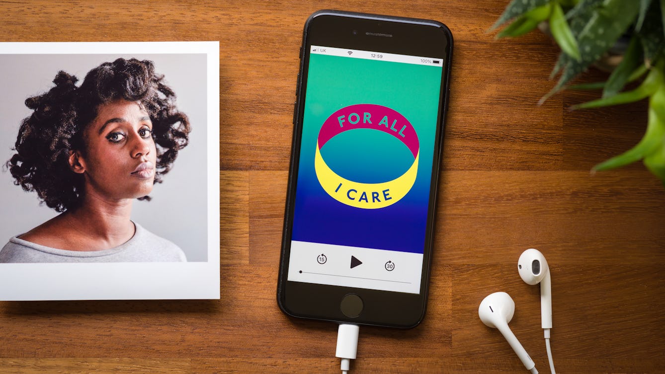 Photograph of a smartphone on a wooden tabletop. Plugged into the phone is a pair of white earbud headphones. On the screen of the phone is a podcast screen with a graphic identity of a coloured ring with the words 'for all I care' written around the ring,. The colours are green, blue, yellow and magenta. To the top right of the image is the green of a pot plant. To the left of the image is a photographic print of a portrait of a woman in a grey top against a grey background, she is looking to camera.