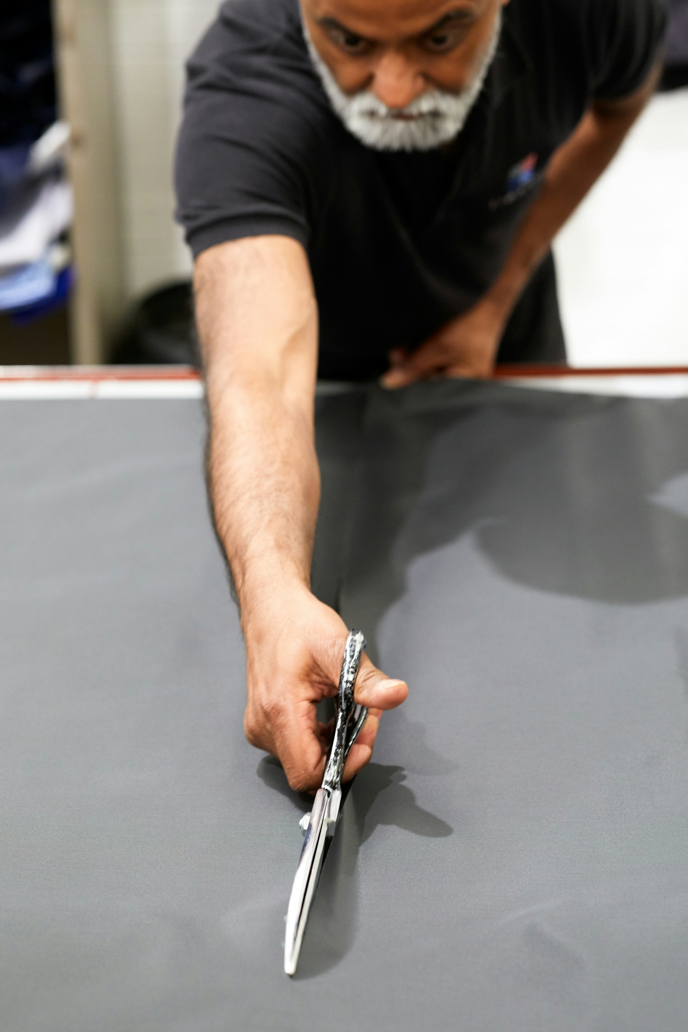 Photograph of a man stretching over a sheet of fabric with a pair of scissors, cutting a straight line away from him.