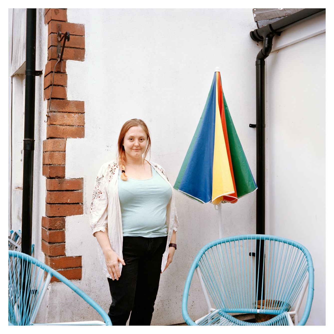 Portrait photograph of Jayne, a woman with blonde and red hair, standing in front of a white painted brick wall. Her hair is in a side braid and she is wearing a light blue t-shirt, a white floral cardigan and black trousers. 

Her hands are by her side and she is looking directly at the camera with a slight smile. There is a colourful patio umbrella next to her and several blue beach chairs. 