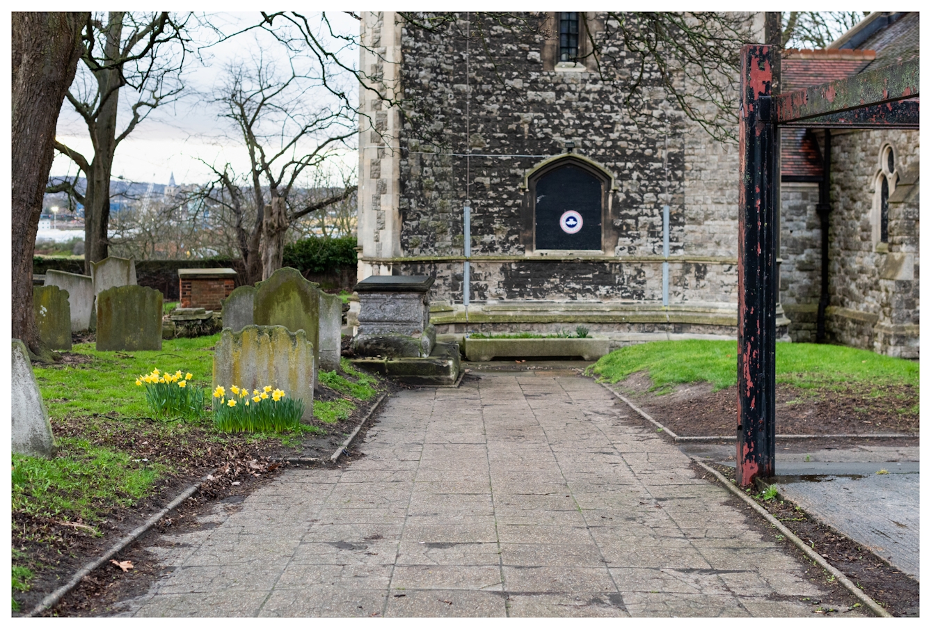 Photograph of a churchyard with the church buildings and leafless trees in the background. In the foreground there are a number of gravestones and grassed areas. To the centre left is small crop of bright yellow daffodils.