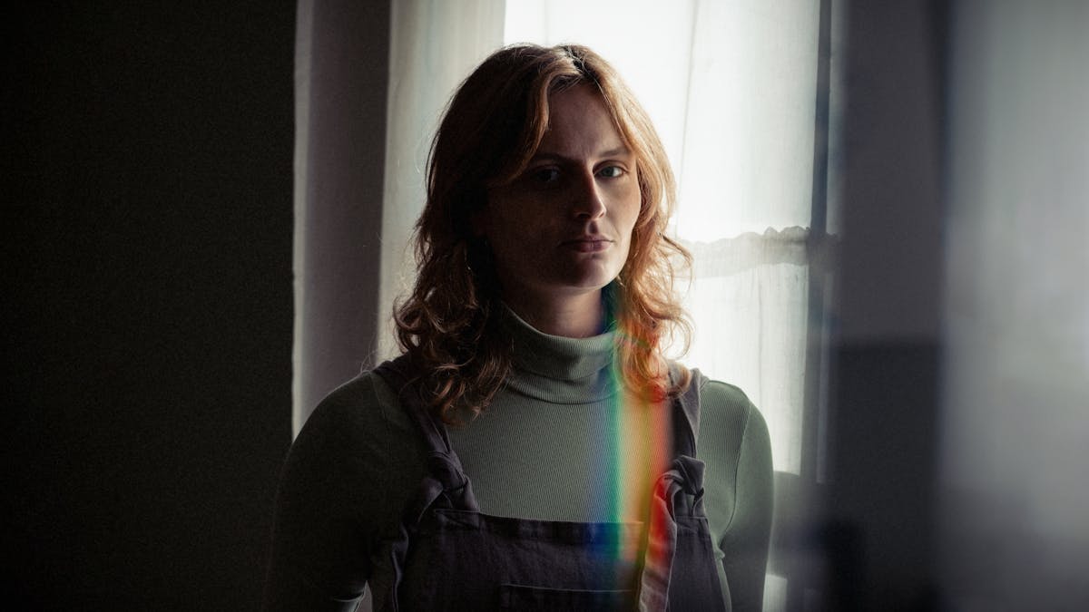A photograph of a woman in front of a window. She is looking directly at the camera and the left hand side of her face is in shadow. She is wearing a green jumper and a pair of blue dungarees. There is a small rainbow beam directly below her face which stretches down to her chest. 