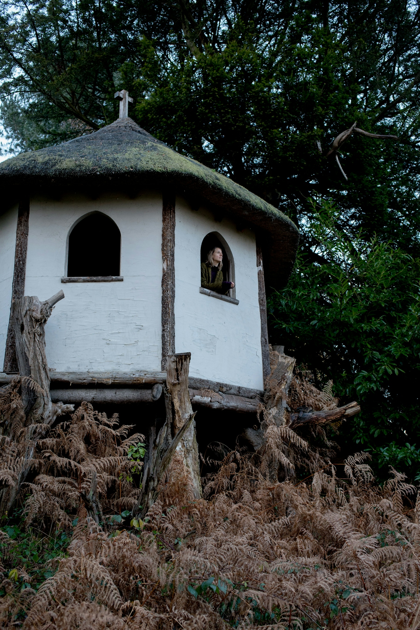 Photograph of a young woman sitting looking out of the window of a hermit's hut, set in woodland.