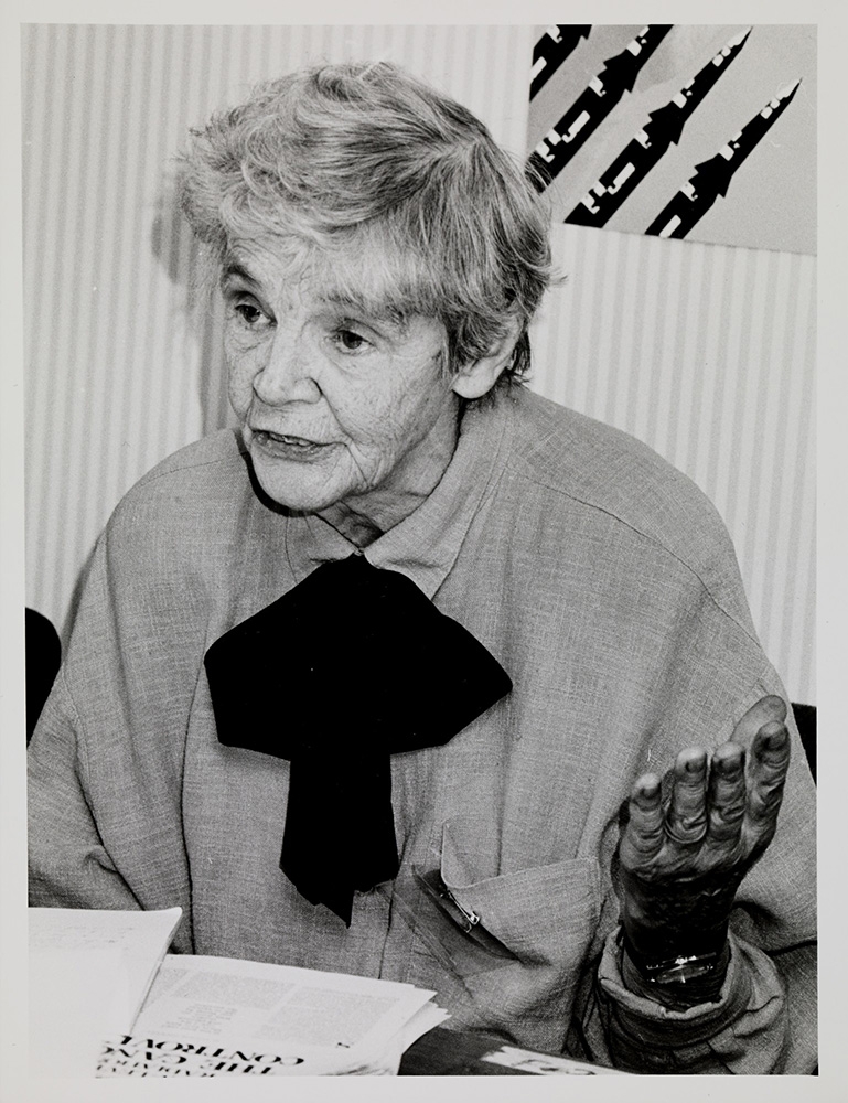 A black and white photo of Alice Stewart, speaking with her hand raised, with a poster of nuclear missiles in the background. 