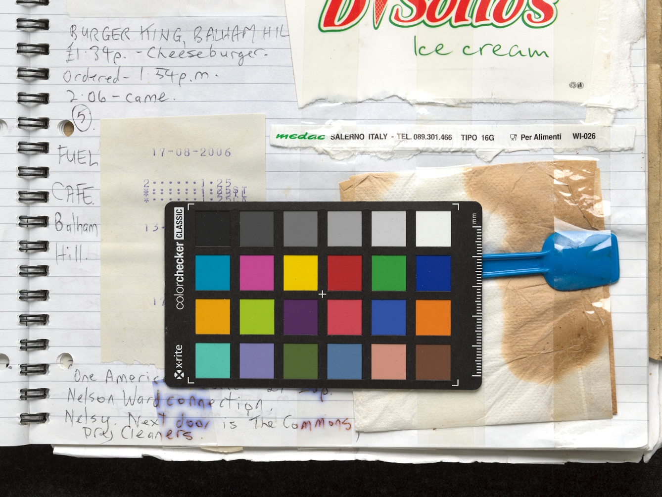 Photograph of a close-up of the lower right hand section of a lines scrapbook. The page contains handwritten notes and receipts, food packaging and napkins have been Sellotaped to the page. Resting on top of the page is a 24 colour patch photographic card, complete with ruled edge in millimetres. The text on the card reads 'ColourChecker'.