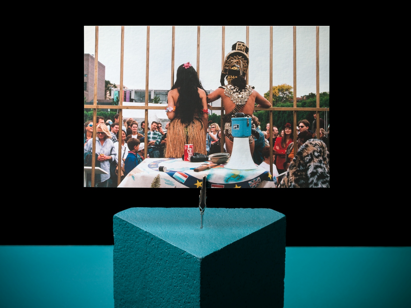 Photograph of a teak triangle on a teal and black background.  A crocodile clip is holding a picture of a performance where two people, a man and a woman, are in costume with their backs to the camera whilst addressing a crowd.  A table of props including a megaphone is behind them.