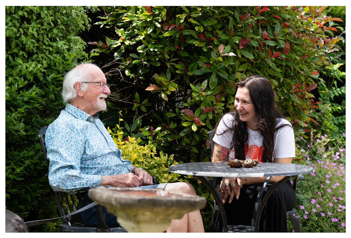 A photograph of two people smiling and laughing with each other in a garden. On the left is an elderly man wearing glasses with white hair and a white goatee beard. He is wearing a shirt covered with small blue flowers. On the right is a young woman with long dark hair. She is wearing a white t-shirt with a red printed design on the front, there are tattoos visible on each arm. Behind both people are large bushes with variegated leaves. 