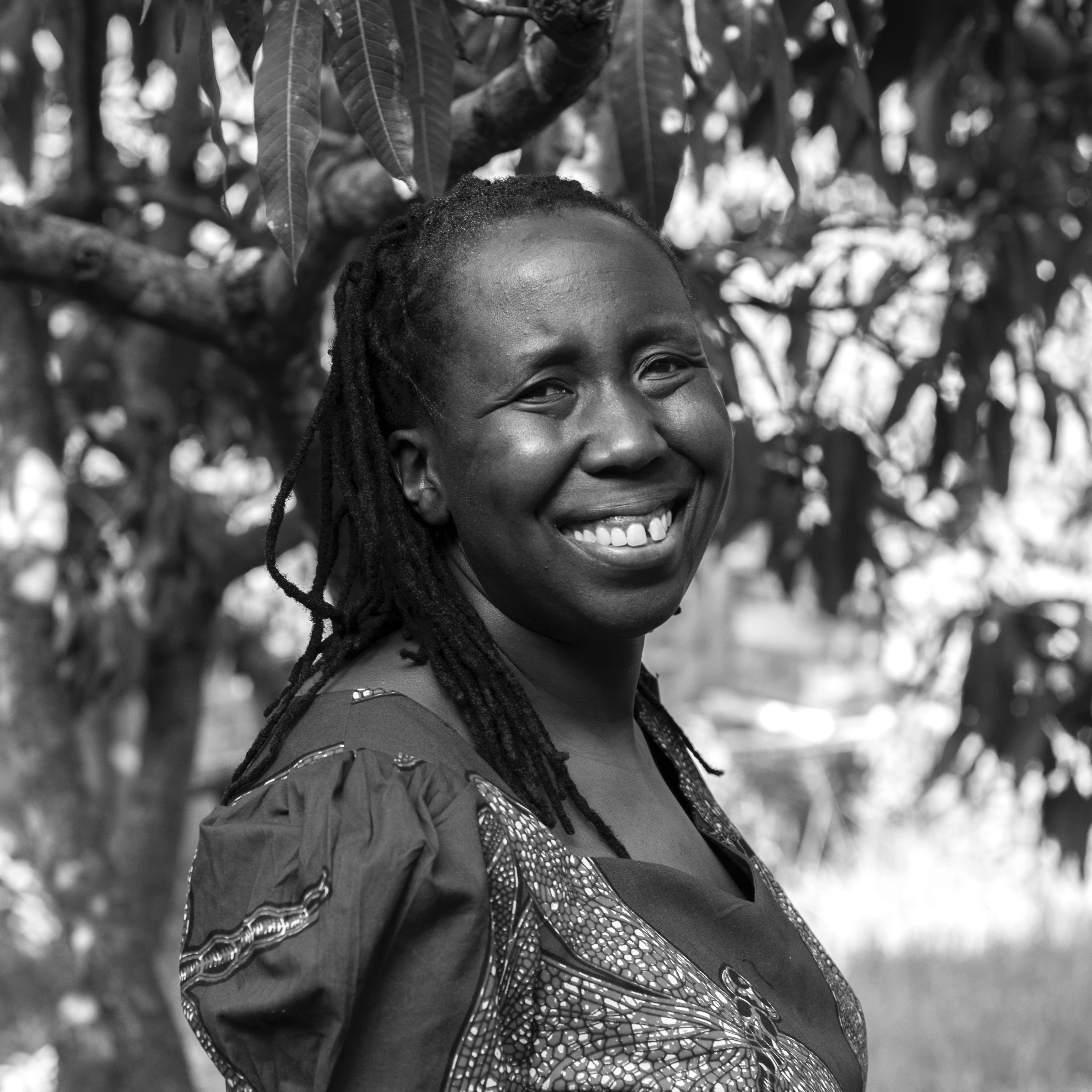 Photograph of a smiling woman with long braids, wearing a print dress and standing in front of a tree.