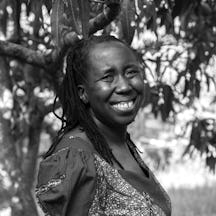 Photograph of a smiling woman with long braids, wearing a print dress and standing in front of a tree.