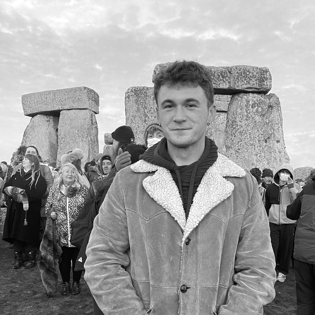 Photograph of a man with short hair, in a warm jacket, standing in front of a crowd of people, behind them are the trilithons of Stonehenge.