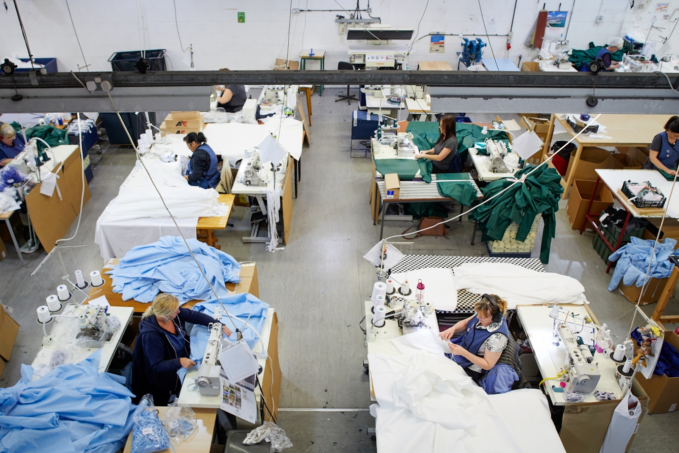 Photograph of a series of workstations within a clothing manufacturing workshop. The viewpoint is from a high walkway which looks down on the workstations. At each workstation is a woman working at a sewing machine, surrounded by white, green and light blue fabric.
