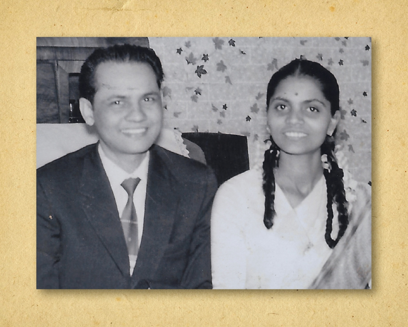 Photograph of a black and white photographic print, resting on a brown paper textured background. The print shows a man and a woman sat side by side smiling to camera. They are both smartly dressed, the man in a suit and tie, and the woman in a dress. Behind them is a patterned wallpaper.