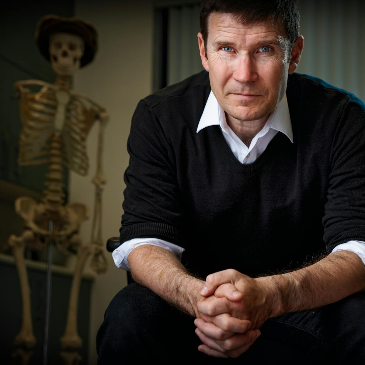 Photograph of a male doctor in his consultation room, leaning forward on his desk chair looking intently into the camera. He is dramatically lit from the side and in the background a skeleton wearing a hat can just be made out.