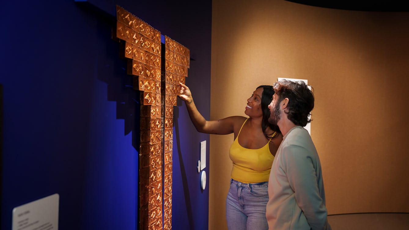 Two people standing in a gallery setting, one is touching an object installation, spotlit against a dark blue wall. The large object is made out of small copper tiles which have hieroglyphics stamped in relief into their surface. The tiles as a whole create a tall straight form rising up the wall which ends with two wing like shapes either side. Either side of the object are information panels and a small framed print.