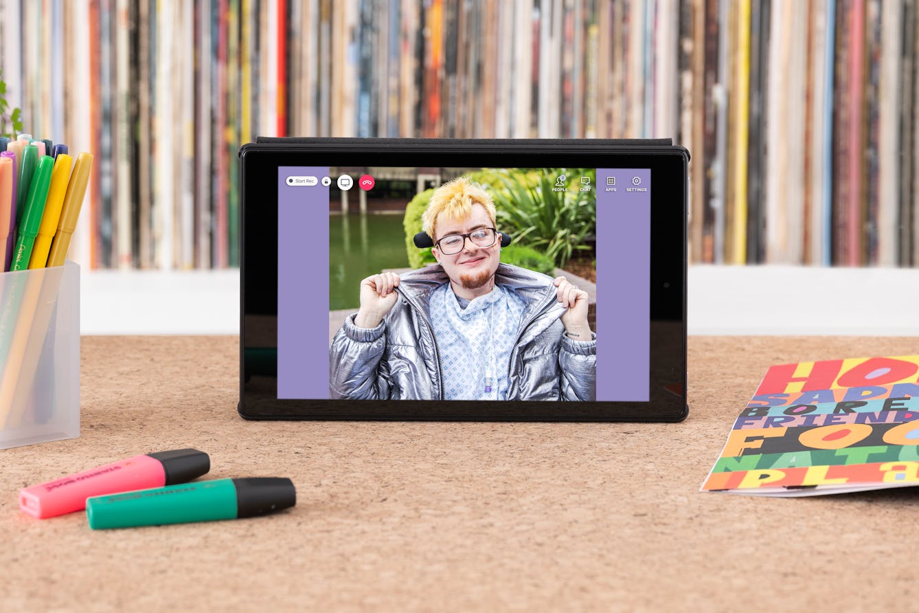 Photograph of a tablet standing on a wooden surface with a vinyl record shelf behind containing the spines of many colourful records. On the tabletop are coloured highlighter pens, a pen pot containing pens and an open magazine showing colourful graphics. A photographic portrait of Jamie Hale is on the tablet screen with video call icons "People", "Chat" and a red telephone icon. 