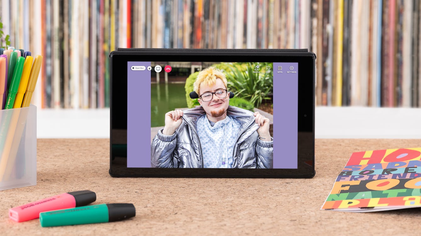 Photograph of a tablet standing on a wooden surface with a vinyl record shelf behind containing the spines of many colourful records. On the tabletop are coloured highlighter pens, a pen pot containing pens and an open magazine showing colourful graphics. A photographic portrait of Jamie Hale is on the tablet screen with video call icons "People", "Chat" and a red telephone icon. 