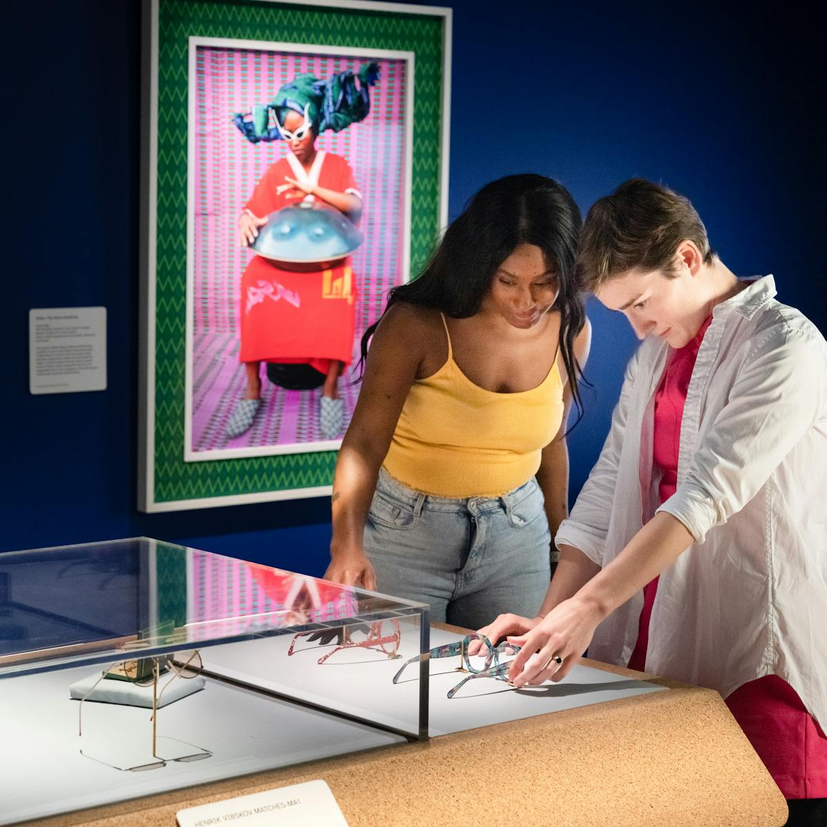 Photograph of two people exploring an exhibition. They are stood side by side at a display case which contains pairs of spectacles. Outside of the case are two pairs of spectacles which the visitors are able to touch and explore with their hands. The rest of the gallery behind them is dark blue, except for a spotlit colourful framed photograph on the wall showing an individual in a bright red dress wearing white rimmed sun glasses with a large domed metal object on their lap.