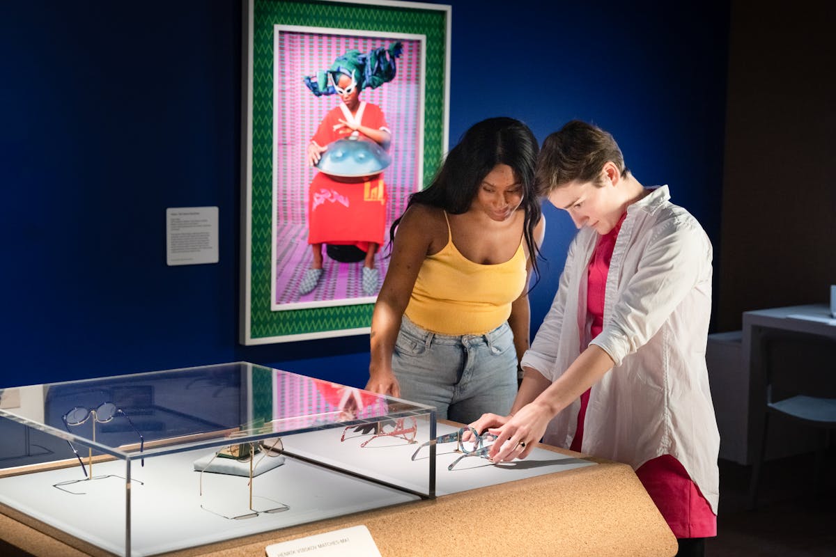 Photograph of two people exploring an exhibition. They are stood side by side at a display case which contains pairs of spectacles. Outside of the case are two pairs of spectacles which the visitors are able to touch and explore with their hands. The rest of the gallery behind them is dark blue, except for a spotlit colourful framed photograph on the wall showing an individual in a bright red dress wearing white rimmed sun glasses with a large domed metal object on their lap.