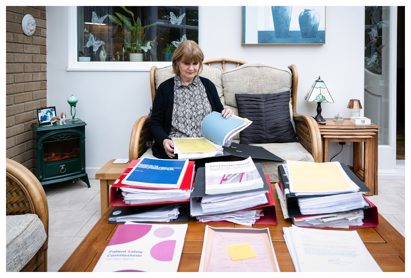 Photograph of Marie Lyon sat on a sofa in her conservatory. She is leafing through a lever arch file on he lap, full of documents. In front of her on the coffee table are 3 piles of lever arch files, papers and printed reports.