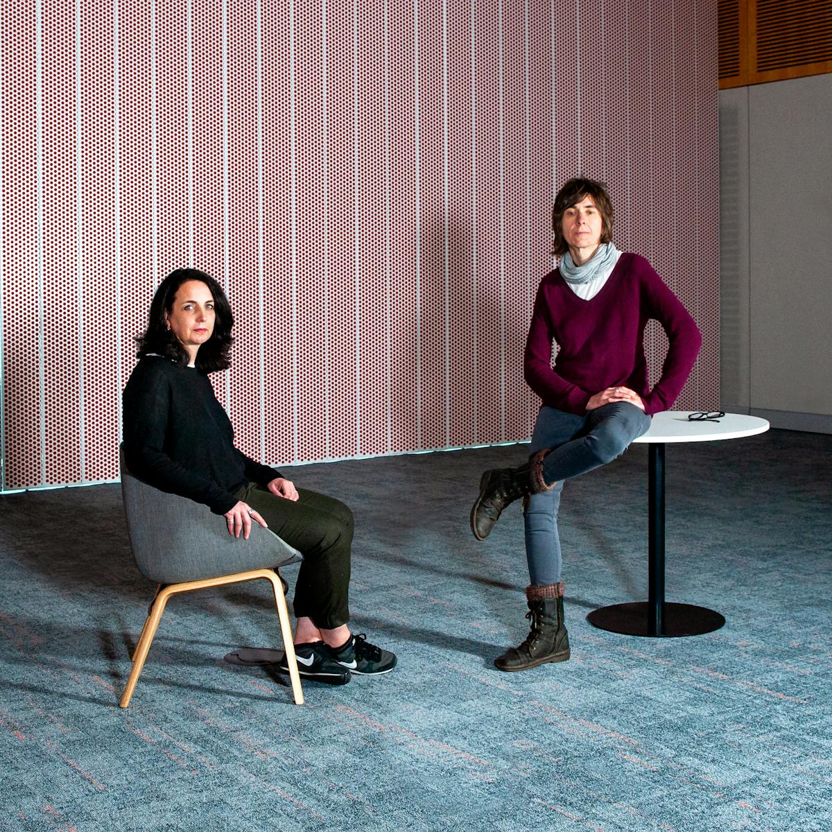 Photographic full length portrait of two women in a meeting room with blue patterned carpet and a red and white spot patterned wall. One woman is seated, sideways to camera looking over her shoulder at the lens. The other woman in sitting on a circular table looking to camera, one leg crossed over the other. Her glasses are folded on the table top beside her.
