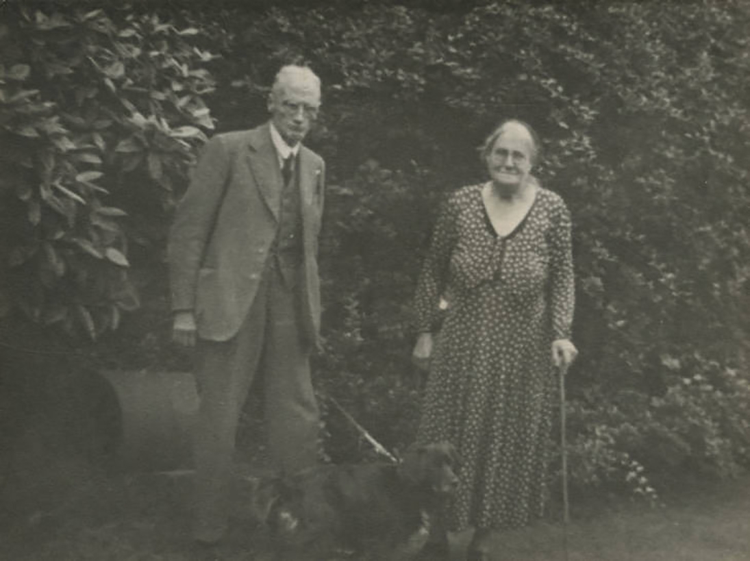 Black and white picture of a man holding a black dog on a lead, and a woman in a spotted dress with a walking stick.