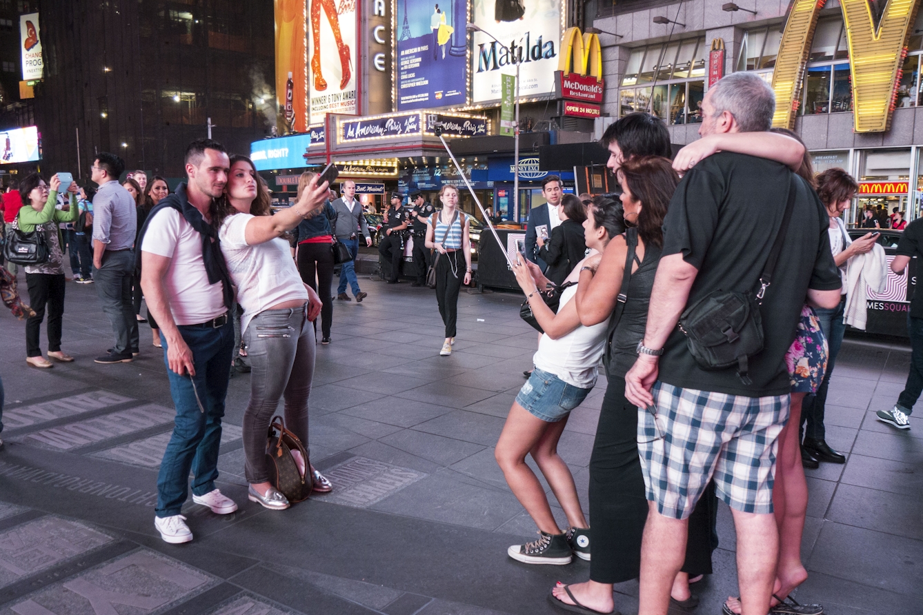 Colour photograph of two groups facing one another whilst taking selfies. On the left a man smiles and a woman pouts whilst leaning backwards to fit into the picture, on the right a woman holds a selfie-stick and leans back towards another two men and two women.
