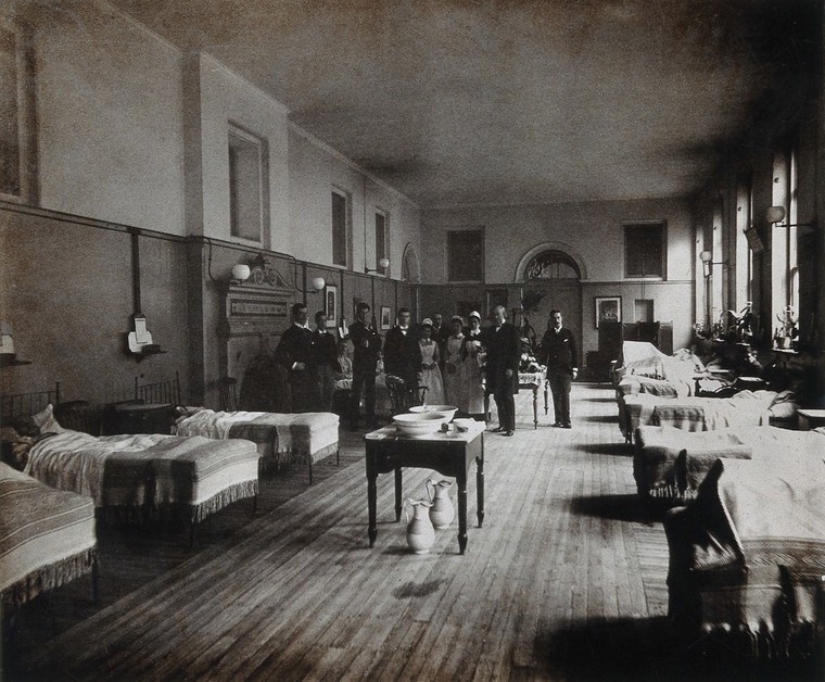 Black and white photograph of a hospital ward with staff posing for the photograph in the middle. The room is lined with beds, which are mostly occupied with patients. There is a table with two large bowls on top and two large jugs underneath in the middle of the room.