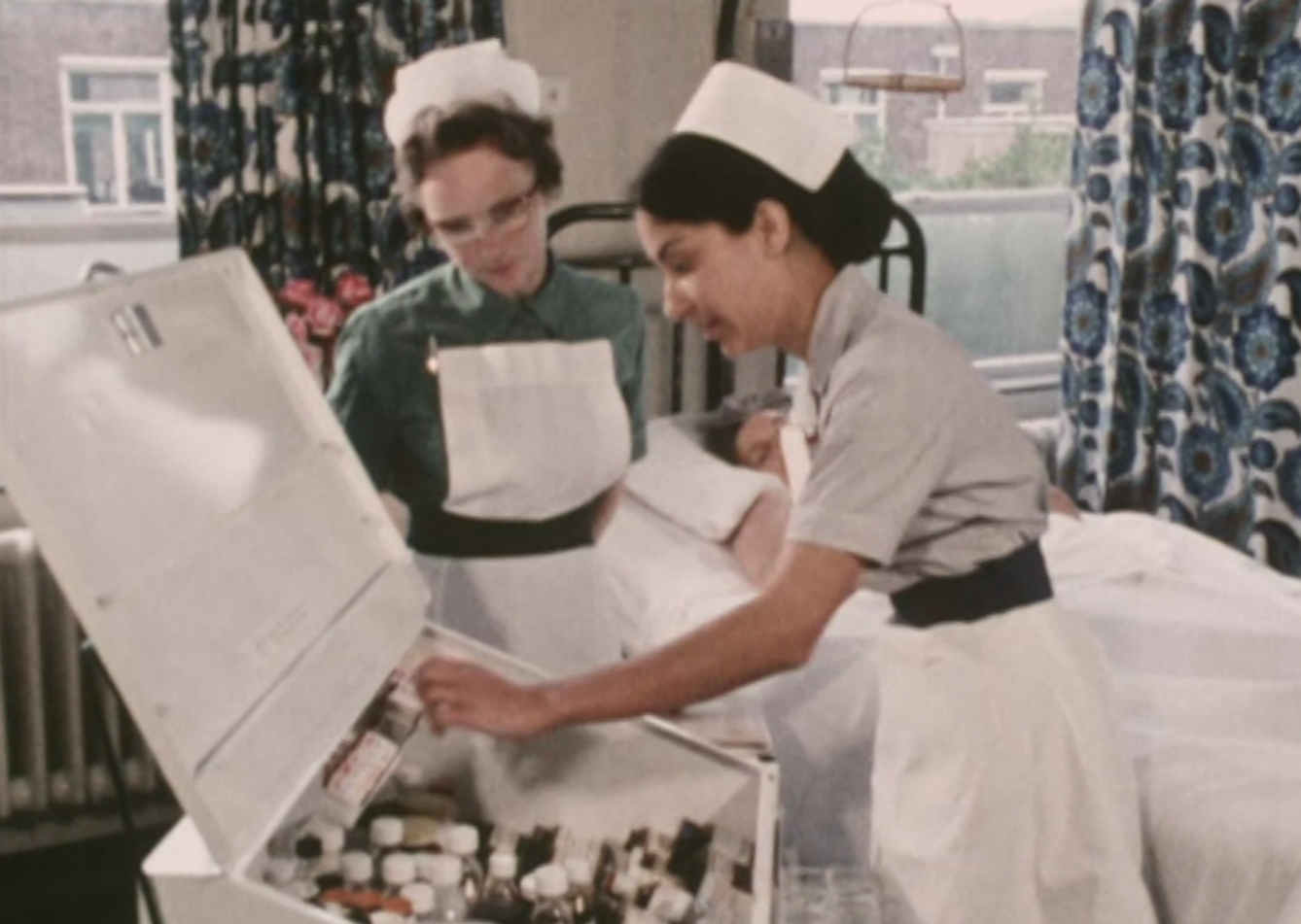 Two nurses leaning over a medical cabinet with a patient lying in a bed in the background.